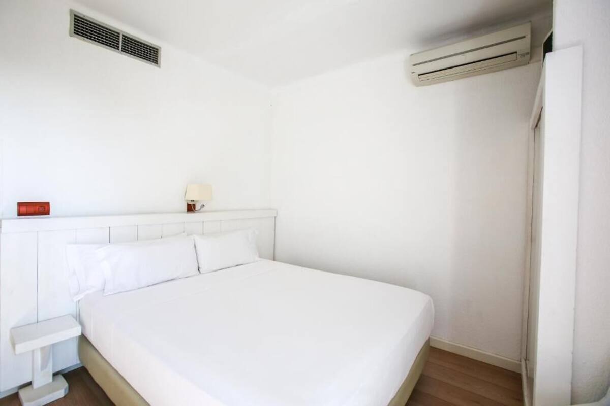 Habitación de hotel blanca e impecable con cama, mesita de noche, lámpara y aire acondicionado.