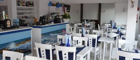 A restaurant with white chairs and tables, a counter with a menu, and a wall with a painting of the ocean.