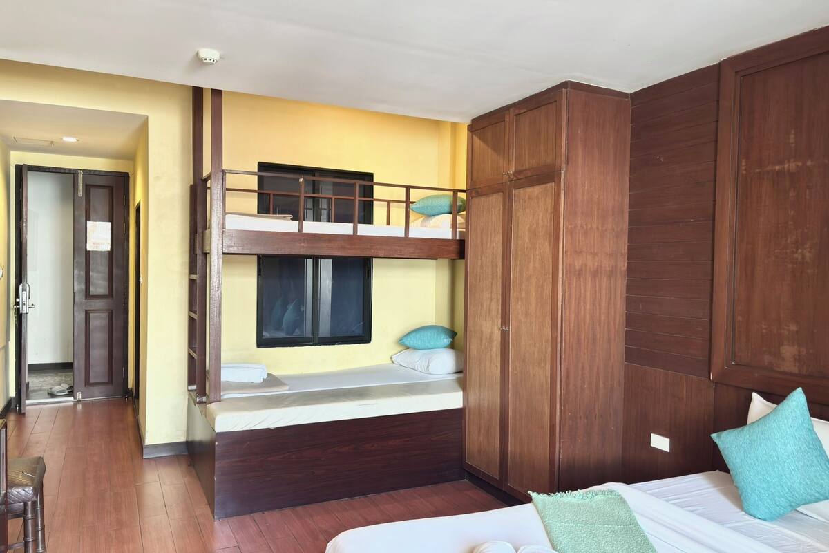 A room with a bunk bed, a large wooden wardrobe, a desk with a chair, and a bed with pillows.