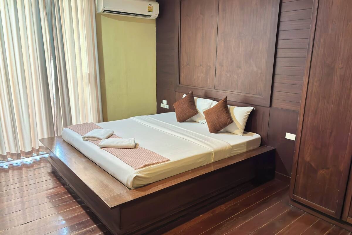 A neatly made bed with pillows and a blanket, a wooden headboard, and an air conditioning unit above it.