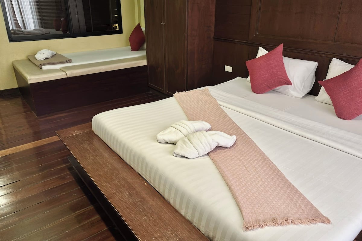 A neatly made bed with white linens, a pink throw blanket, and two white towels. There is a bench with a red pillow and a white towel beside a window with blinds.