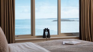 Et soveværelse med et stort vindue med udsigt over havet, en seng og en kikkert på vindueskarmen.