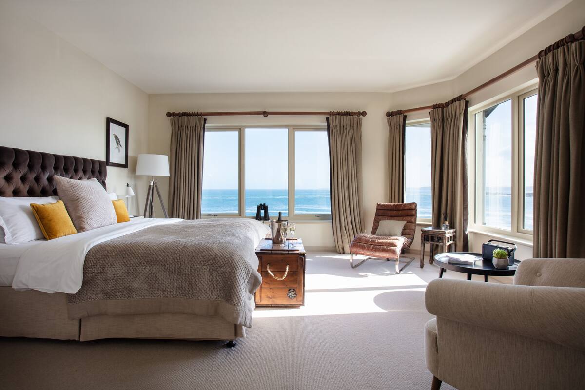 A spacious bedroom with a large bed, a sitting area with a chair, a small table, and a view of the sea.