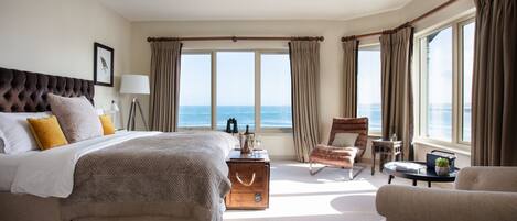 A spacious bedroom with a large bed, a sitting area with a chair, a small table, and a view of the sea.