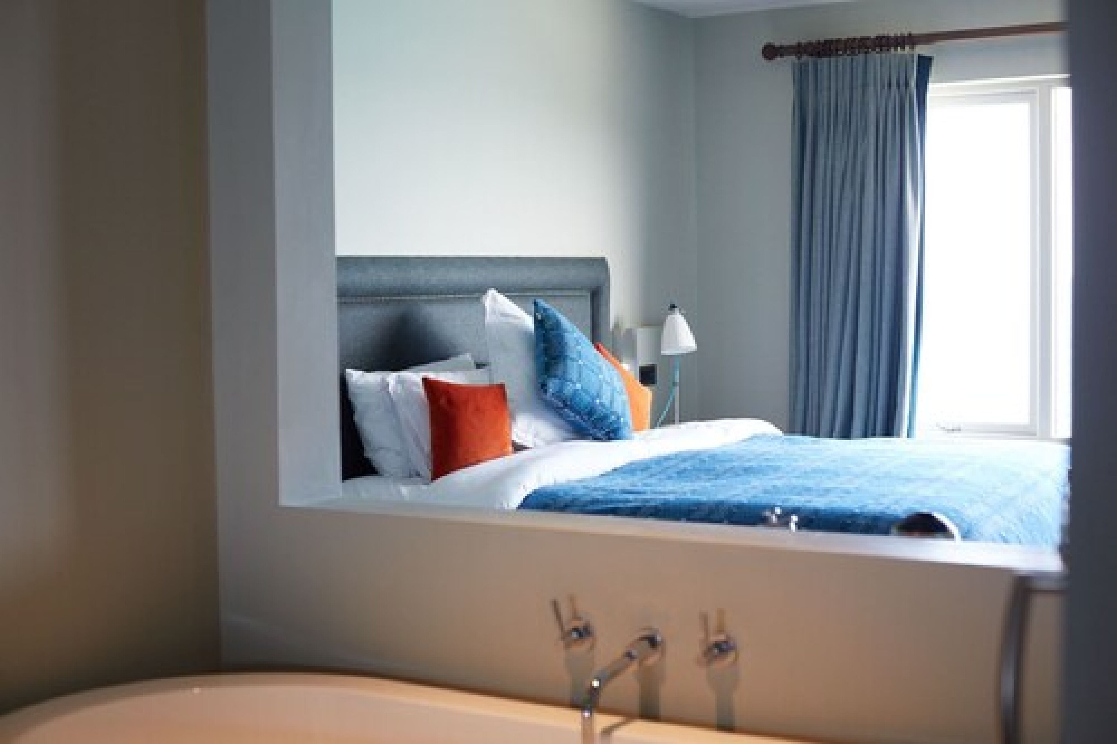A modern hotel room with a large bed, a nightstand, a lamp, and a view of the outside through a window with curtains.