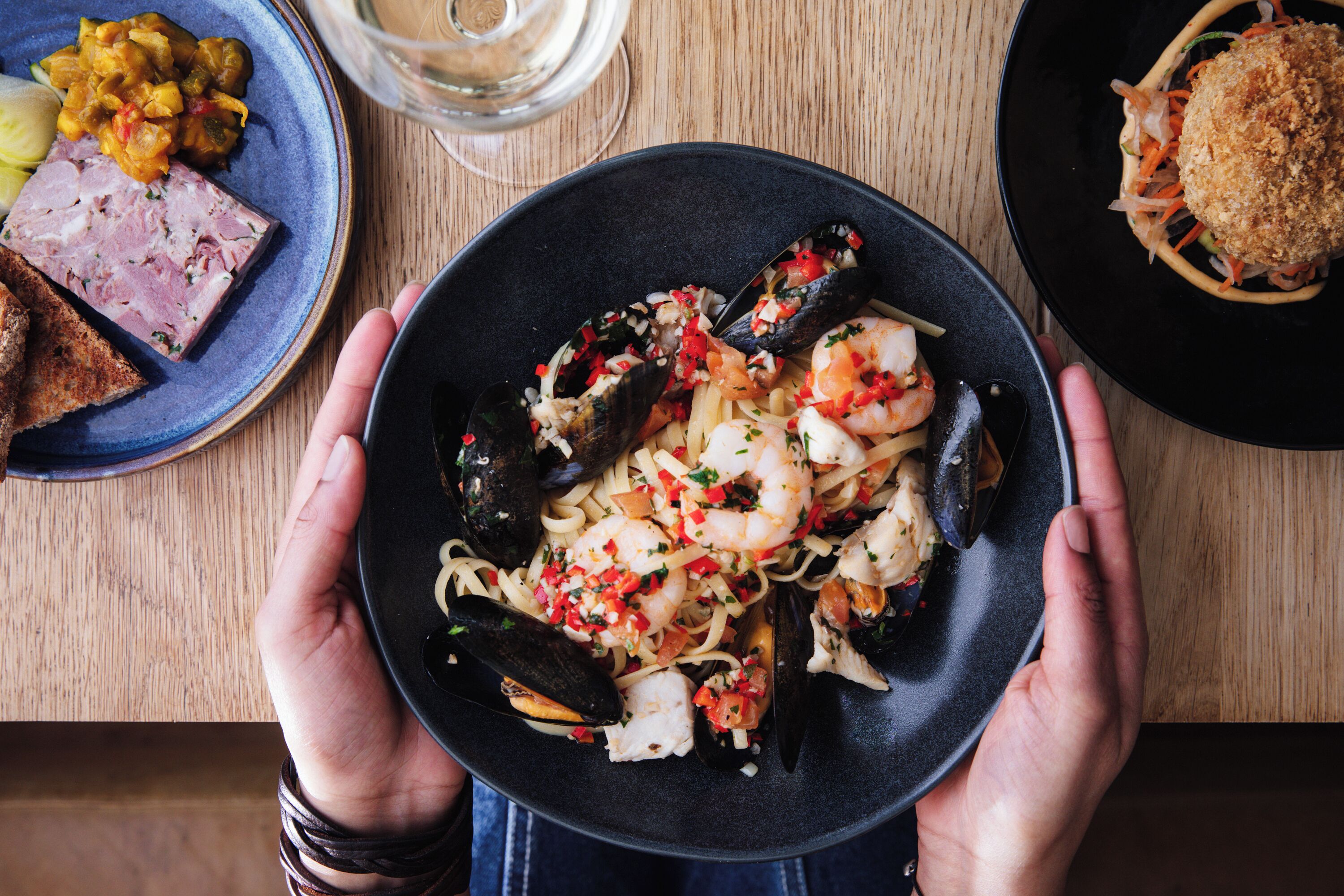 Eine Person hält einen Teller mit Meeresfrüchte-Pasta mit Muscheln und Garnelen, auf dem Tisch sind weitere Gerichte zu sehen.