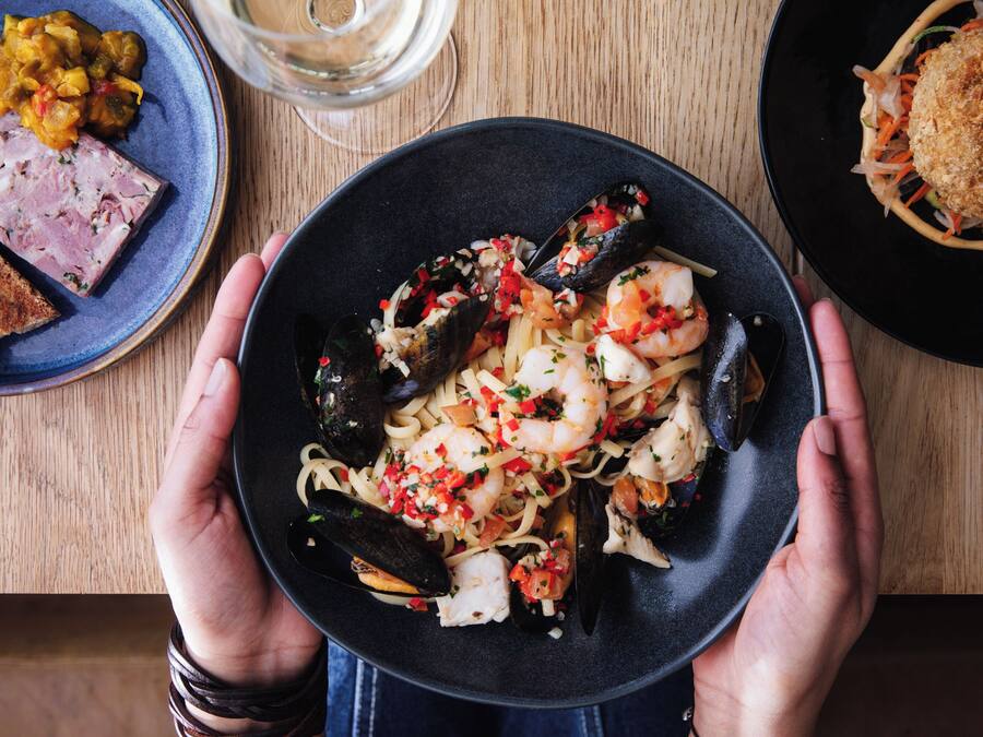 Eine Person hält einen Teller mit Meeresfrüchte-Pasta mit Muscheln und Garnelen, auf dem Tisch sind weitere Gerichte zu sehen.