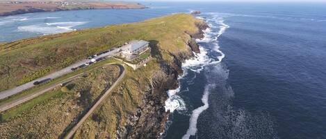 An aerial view of a coastal area with a prominent cliff, a road curving along the coastline, and a building with a flat roof near the cliff base.