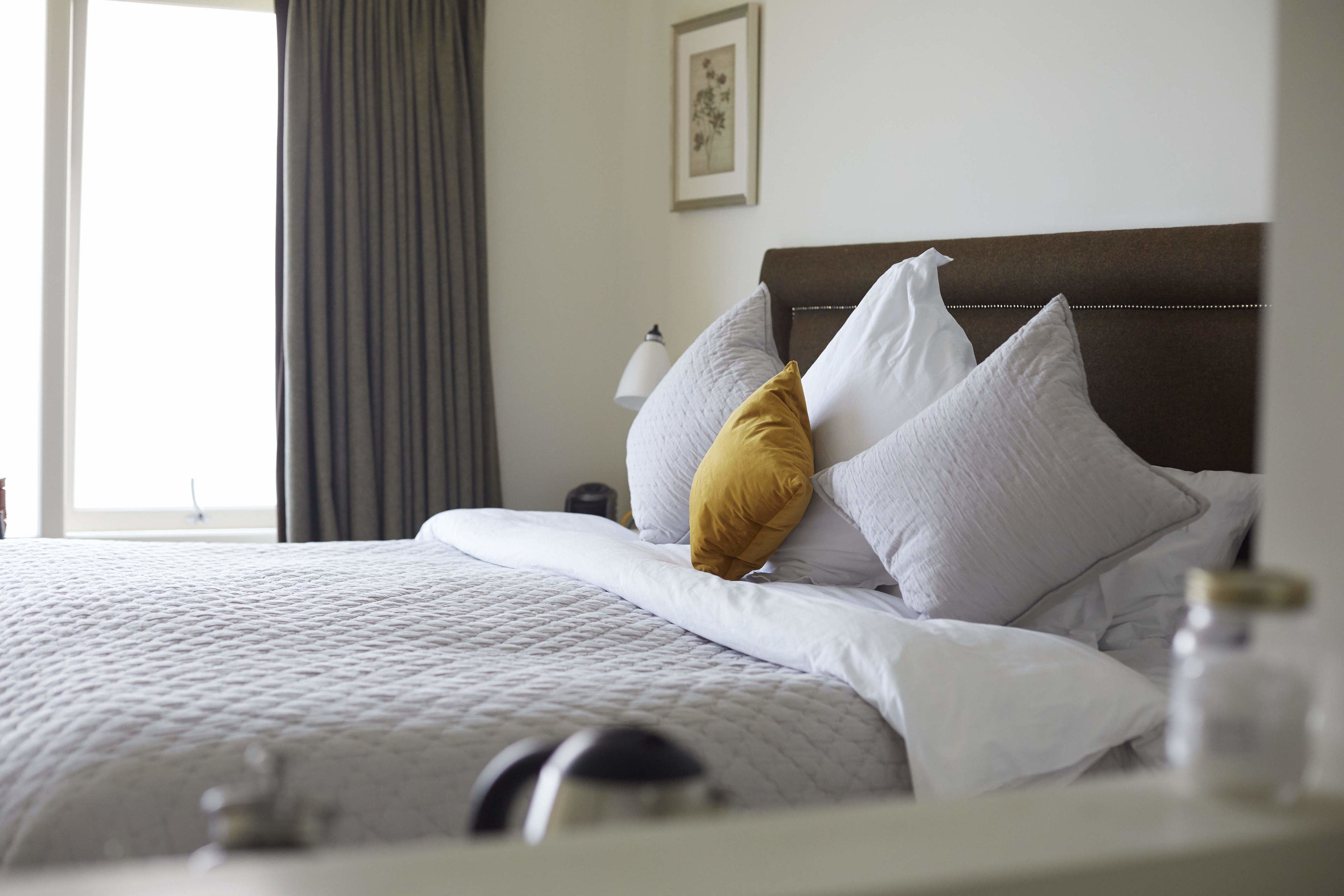 A neatly made bed with white and mustard pillows, a brown headboard, and a bedside lamp.