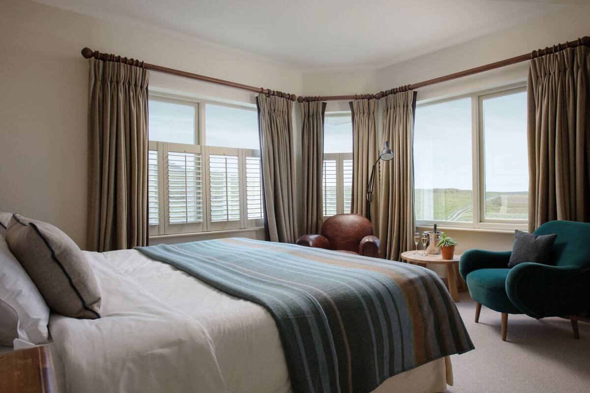 A hotel room with a large bed, a chair, a small table with a lamp, and a view of the outdoors through the windows.