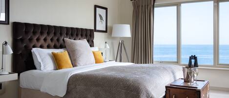 A bedroom with a large bed, a bedside lamp, a nightstand, a wooden chest, and a view of the ocean through a window.