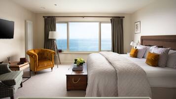 Habitación de hotel con una cama grande, un rincón de descanso con una silla, un escritorio y vista al mar a través de un ventanal amplio.