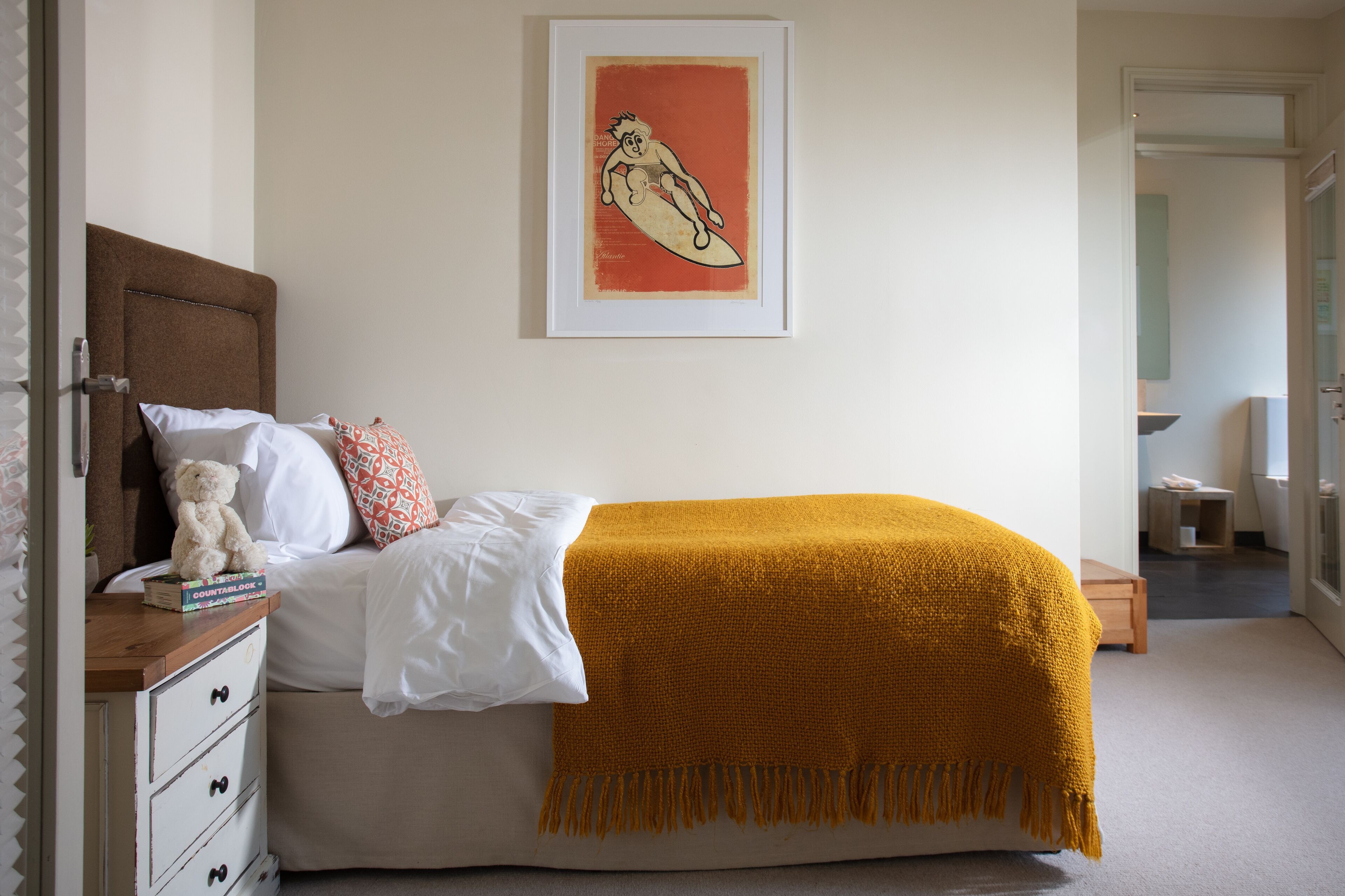 A bedroom with a bed, a bedside table with a teddy bear, a framed picture on the wall, and a door leading to another room.