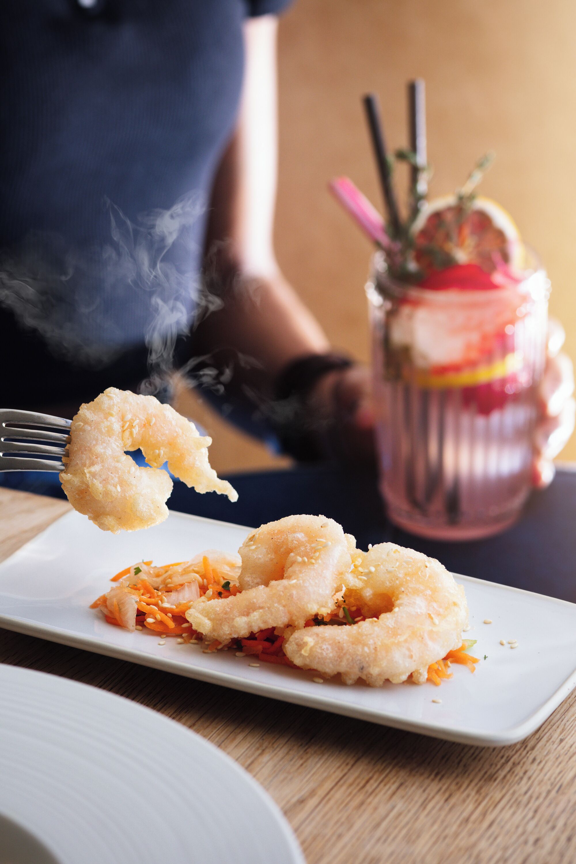 Une assiette de crevettes frites, une fourchette soulevant une crevette, un verre de boisson glacée avec une paille et une tranche d’orange, et une personne floue en arrière-plan.