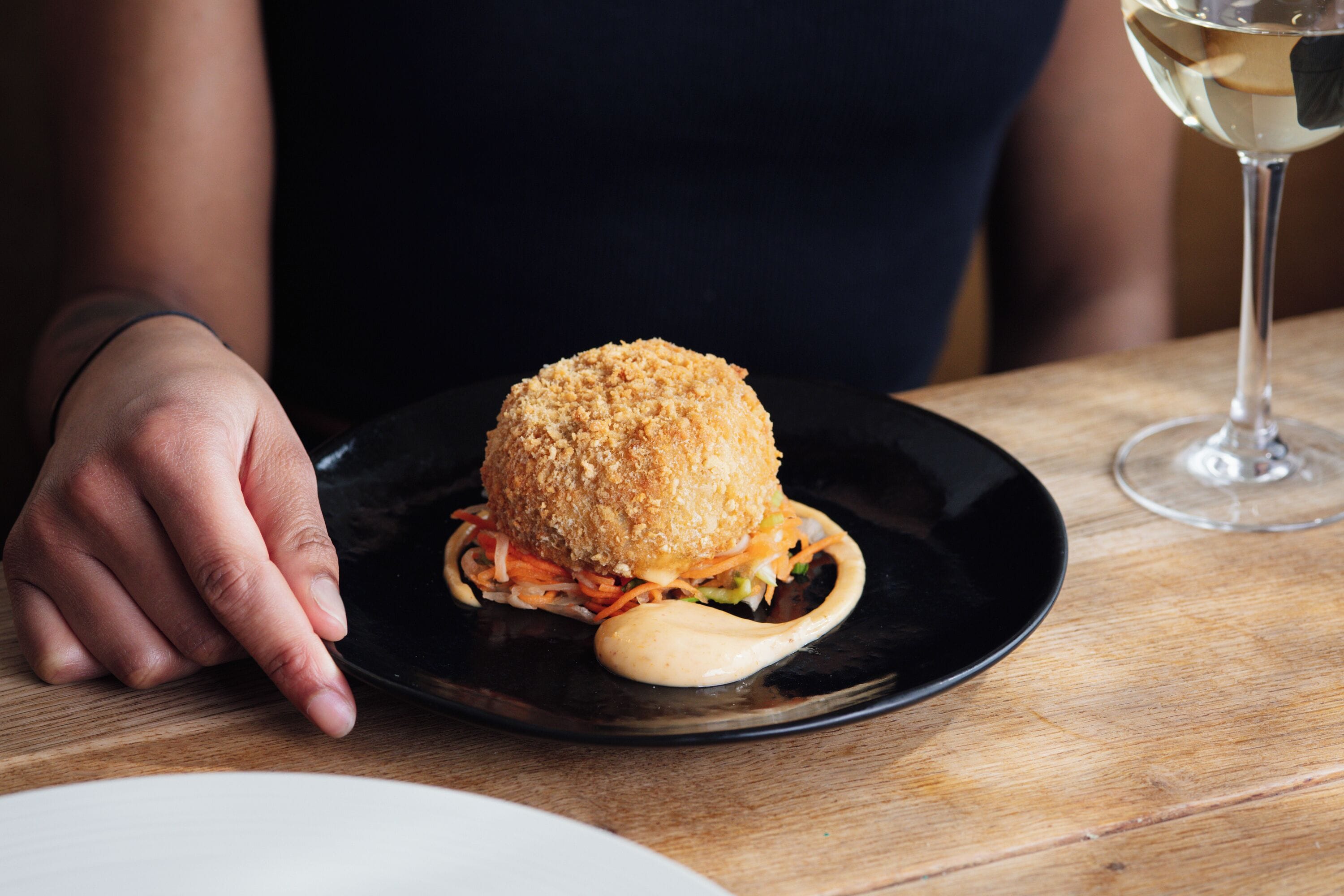 A hand resting on a table with a plate containing a sandwich and a glass of white wine.