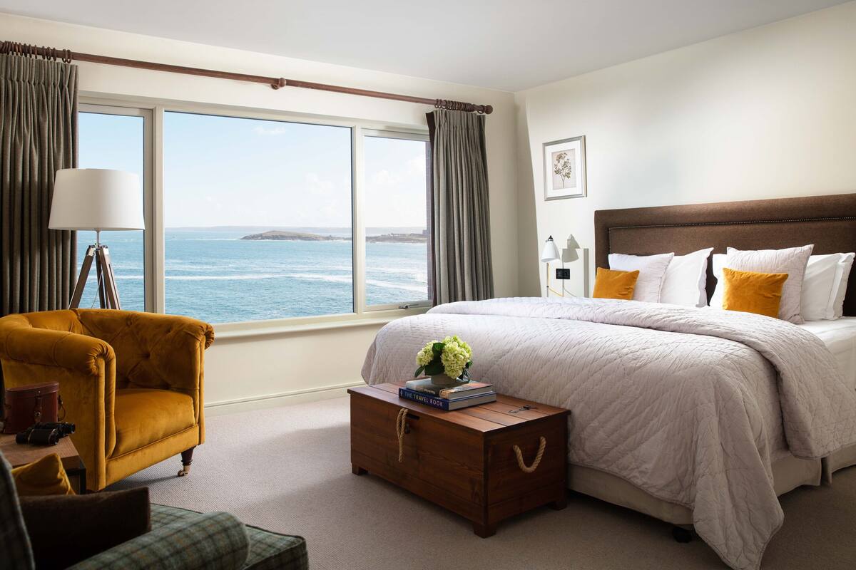 A bedroom with a large window offering a sea view, a bed with white and yellow pillows, a wooden nightstand with a vase and books, and a yellow armchair.