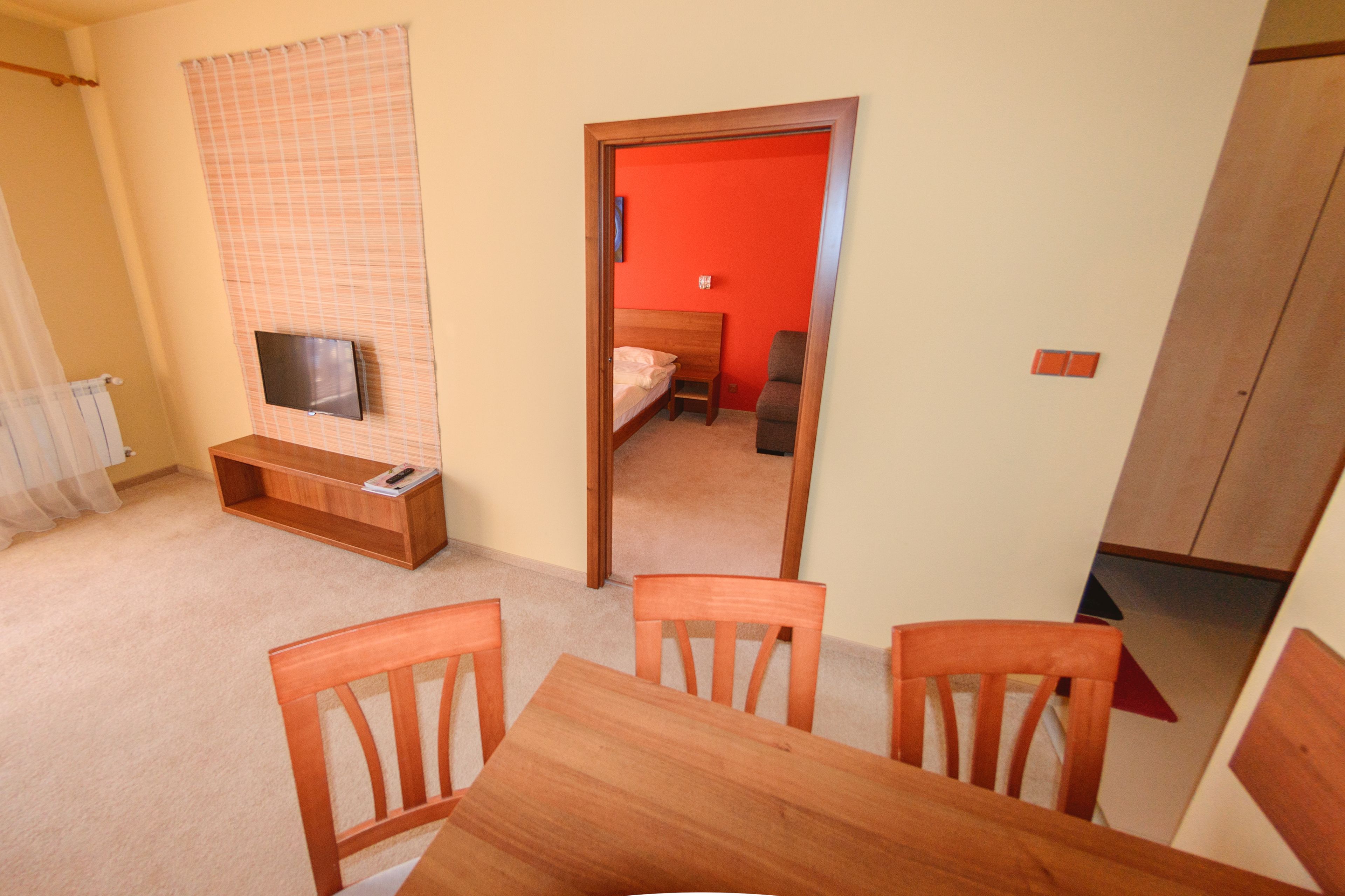 Um quarto com uma mesa de jantar de madeira, cadeiras, uma televisão na parede e uma porta vermelha que dá para outro cômodo.