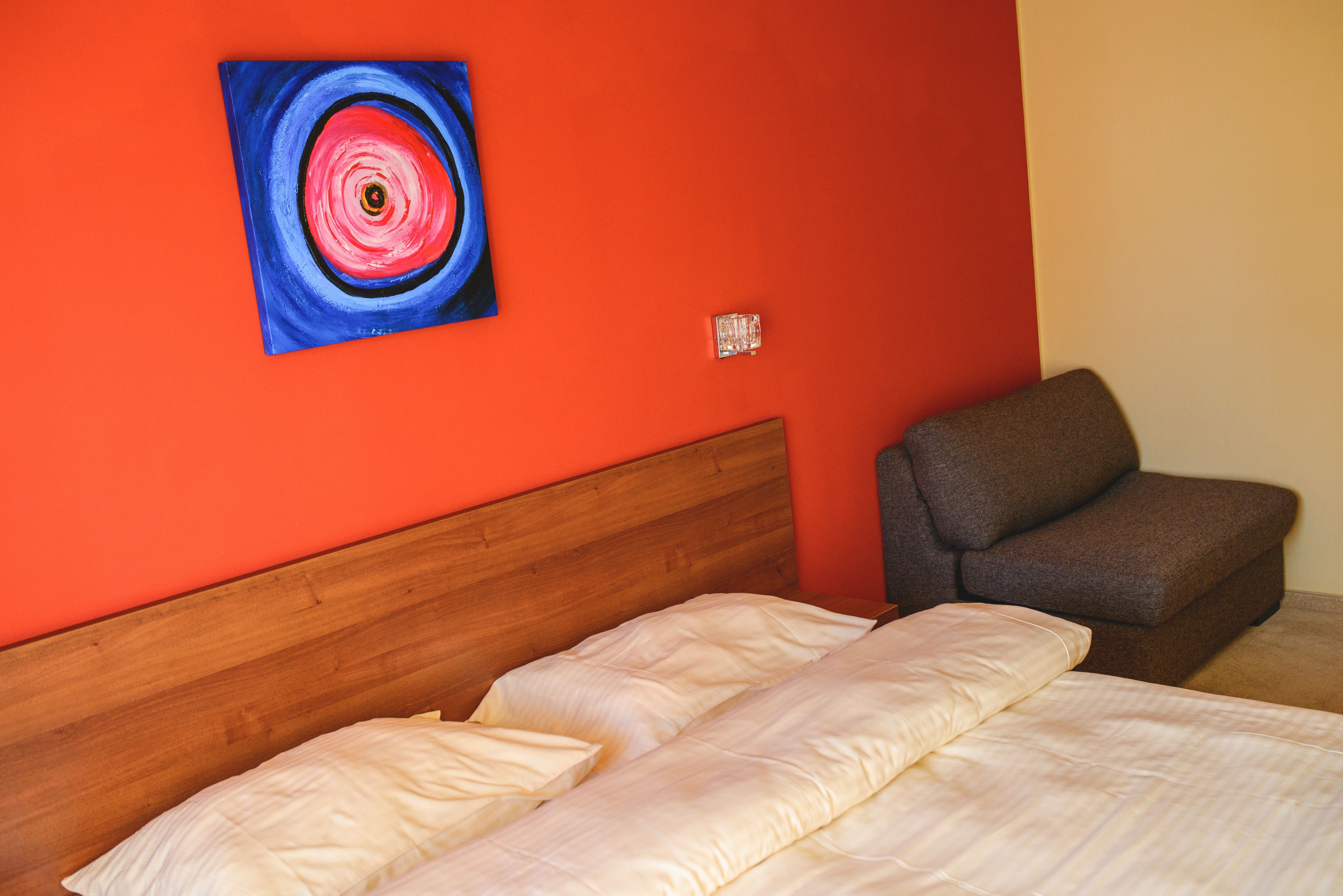 A room with a wooden headboard, a bed with white bedding, a gray armchair, and a red wall with a circular painting.