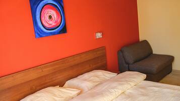 Una habitación con cabecera de madera, una cama con ropa de cama blanca, un sillón gris y una pared roja con un cuadro circular.