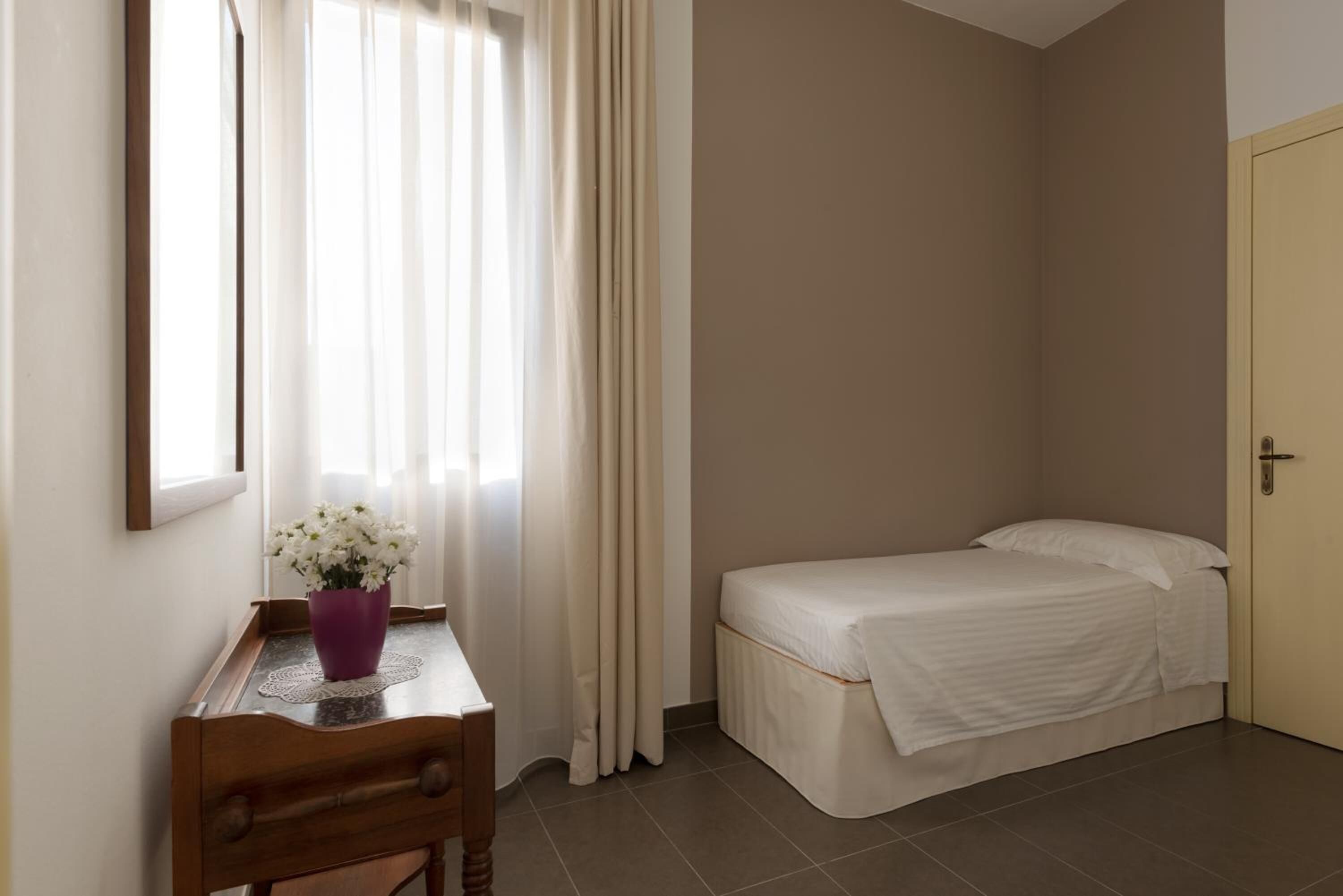 A bedroom with a bed, a desk with a vase of flowers, a window with curtains, and a door.