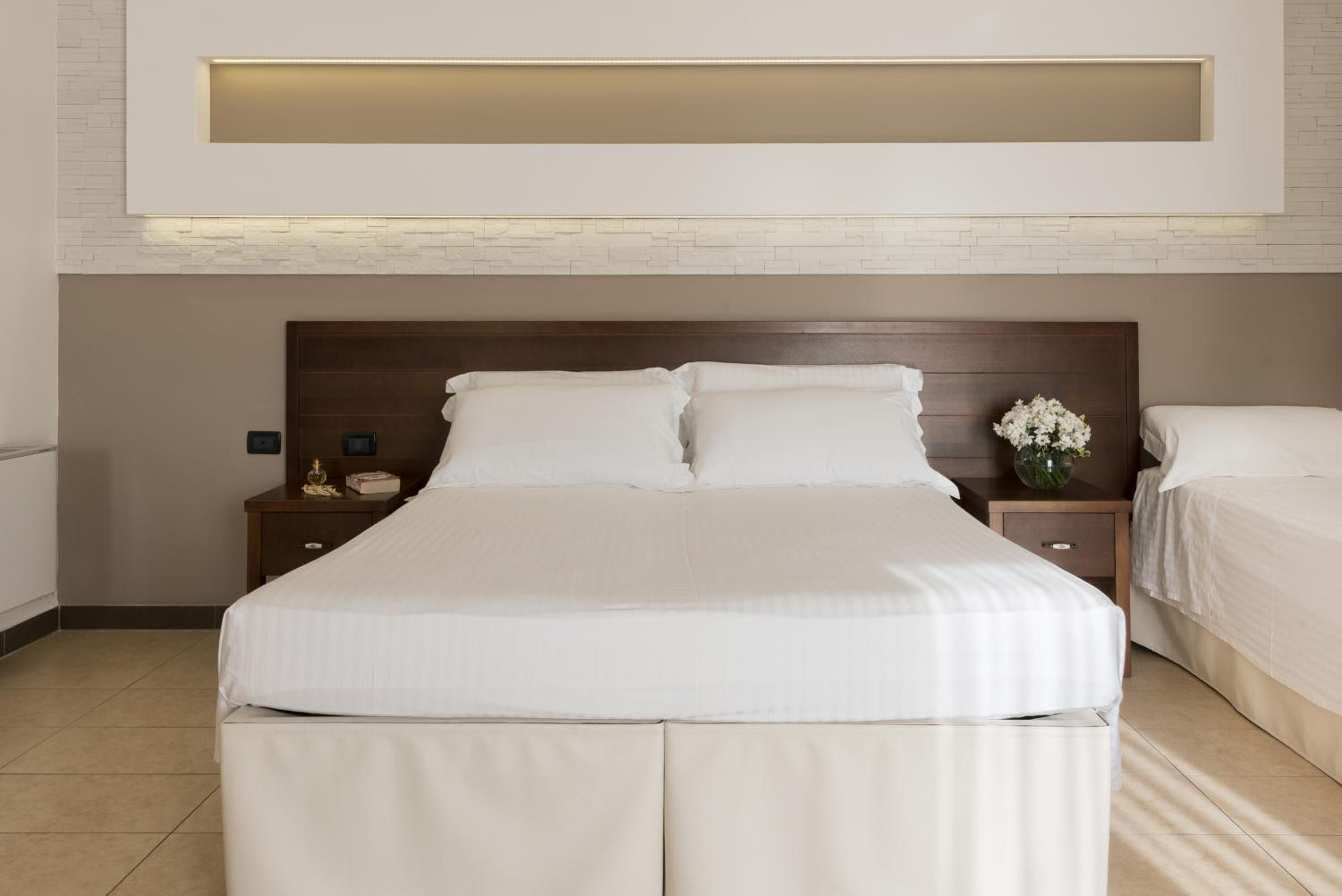Quarto de hotel moderno com uma cama grande, cabeceira de madeira e mesa de cabeceira com um vaso de flores.
