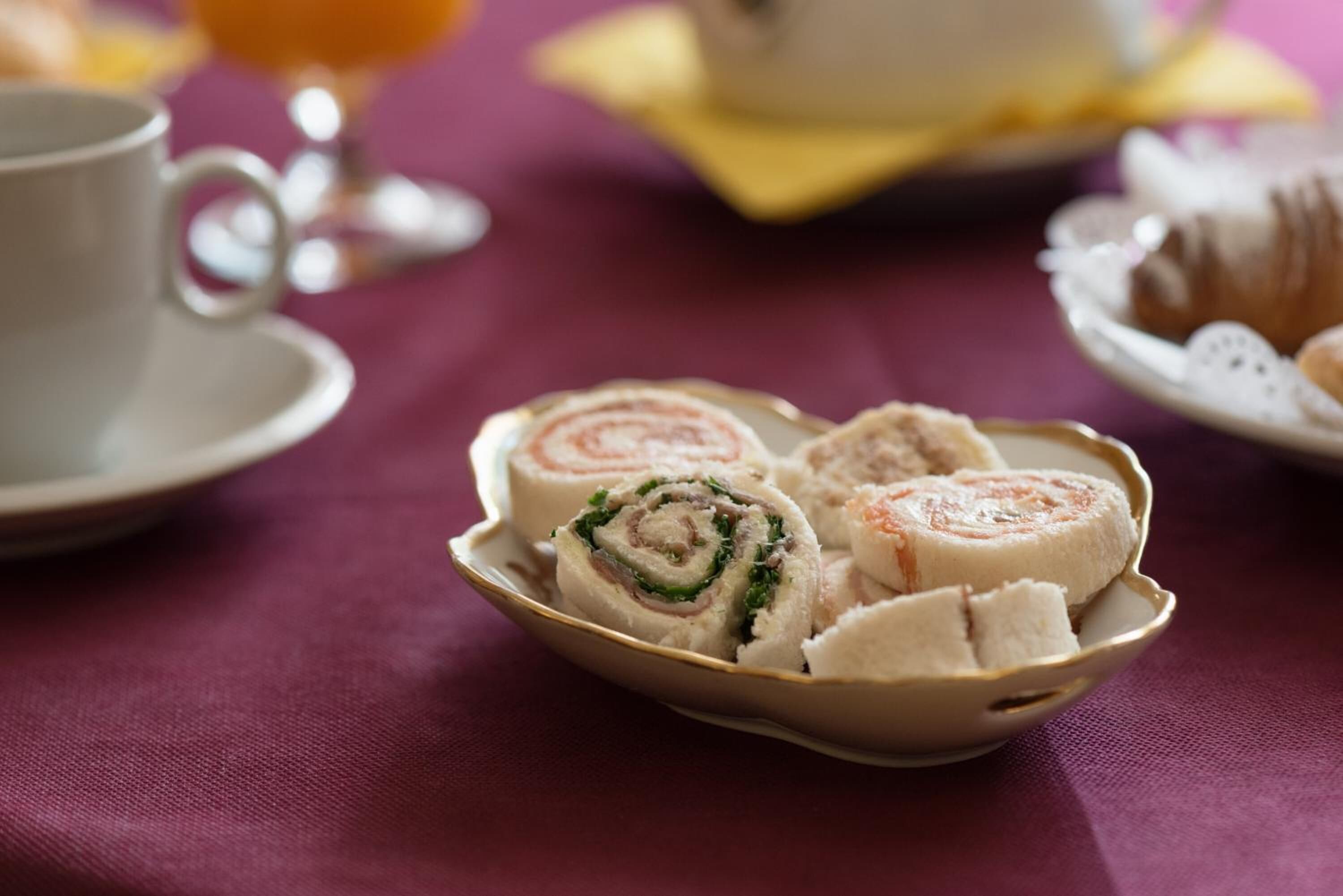 Une assiette de sandwichs coupés en tranches est posée sur une table, avec une tasse et une assiette de pâtisseries en arrière-plan.