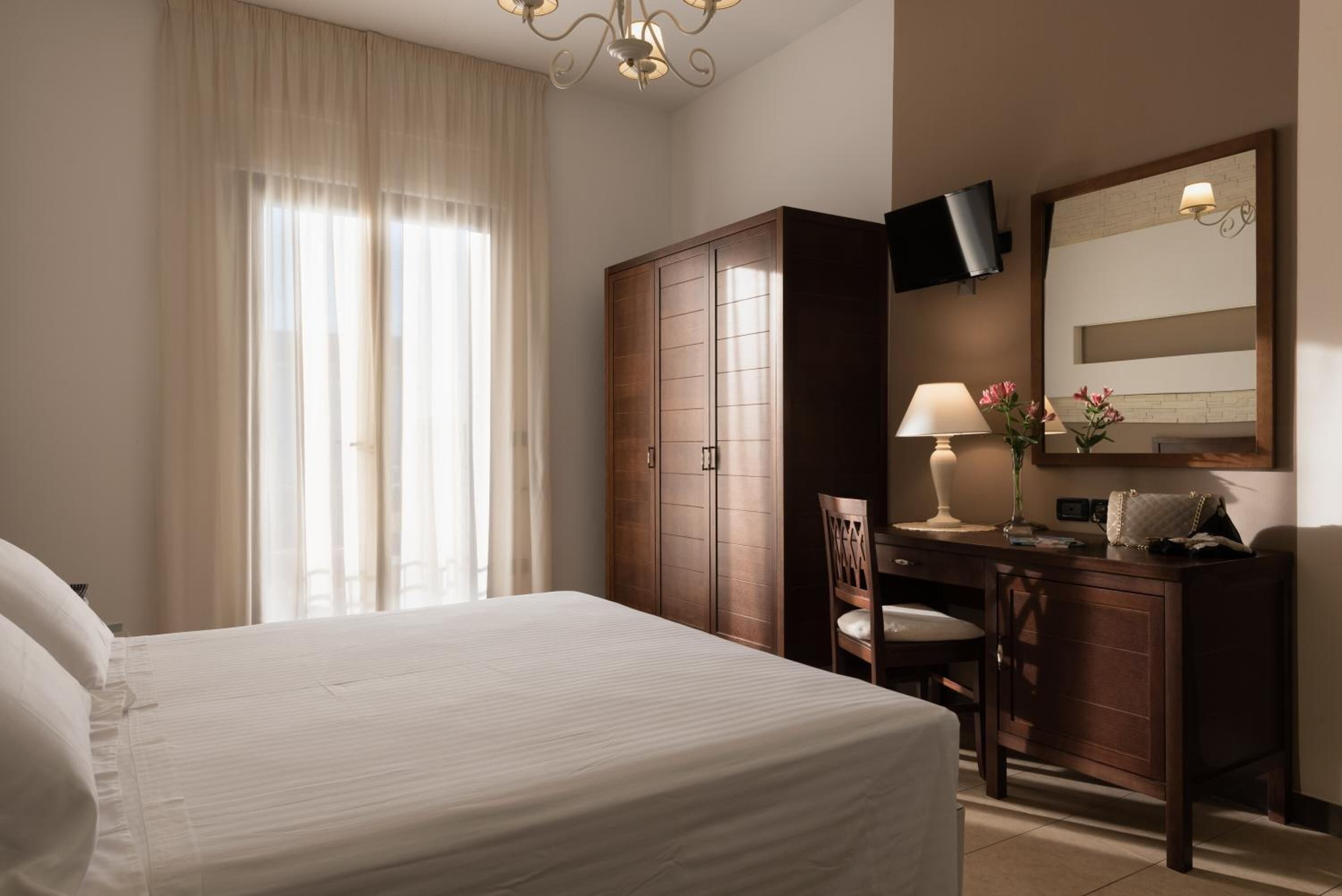 Una camera d'albergo con un letto grande, un armadio in legno, una scrivania con una sedia, una televisione a muro e una finestra con tende.