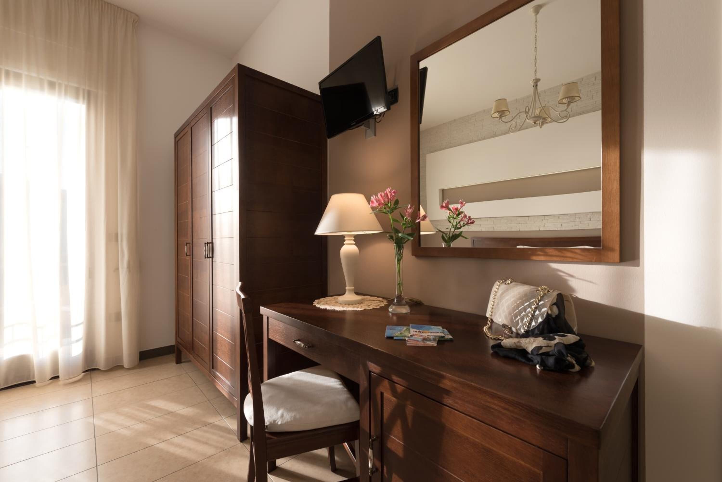 A hotel room with a wooden desk, a chair, a TV mounted on the wall, a mirror, a chandelier, and a window with curtains.