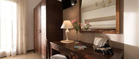 A hotel room with a wooden desk, a chair, a TV mounted on the wall, a mirror, a chandelier, and a window with curtains.