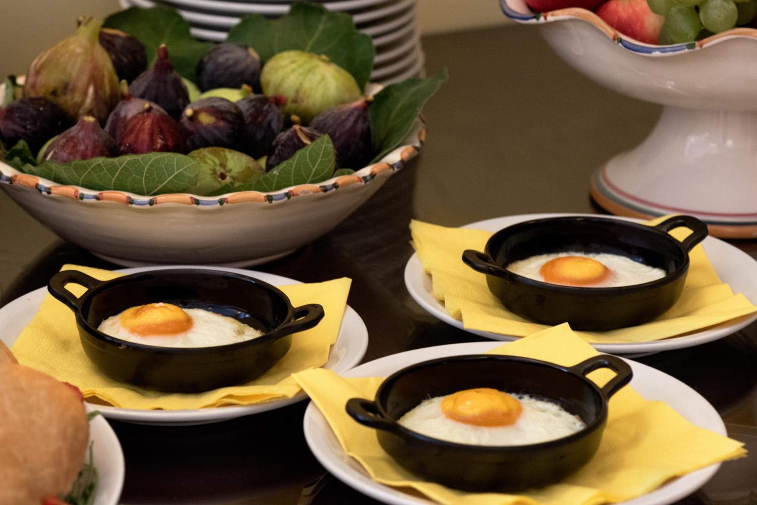 Three black ramekins with eggs on white plates, a bowl of figs and leaves, and a white decorative bowl in the background.