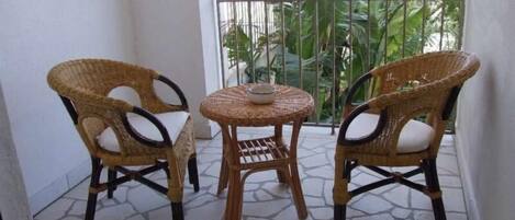 A balcony with two wicker chairs and a small round table, featuring a stone floor and a view of greenery.