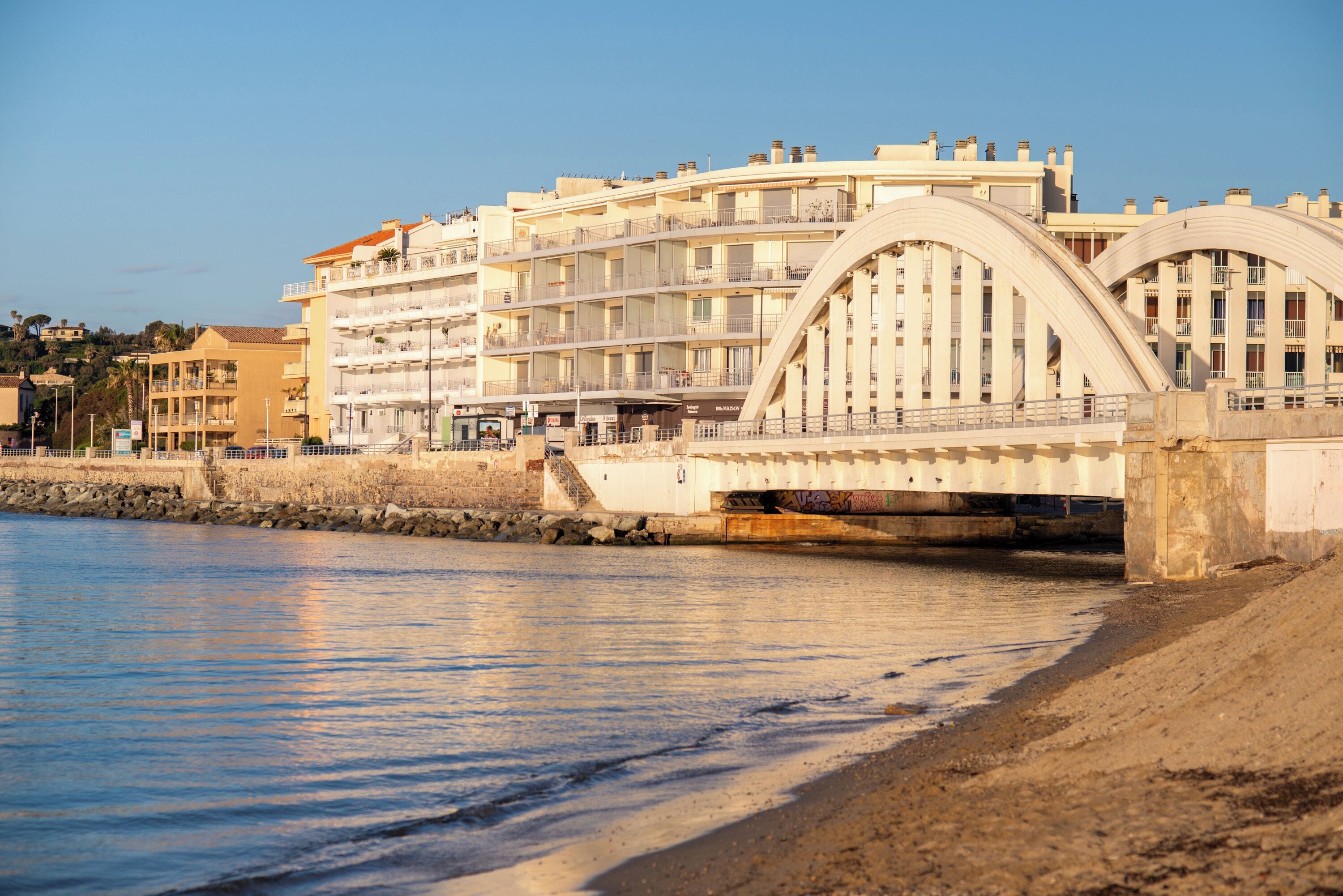 Ein mehrstöckiges Hotel mit Balkonen, ein Strand im Vordergrund und eine Brücke im Hintergrund.