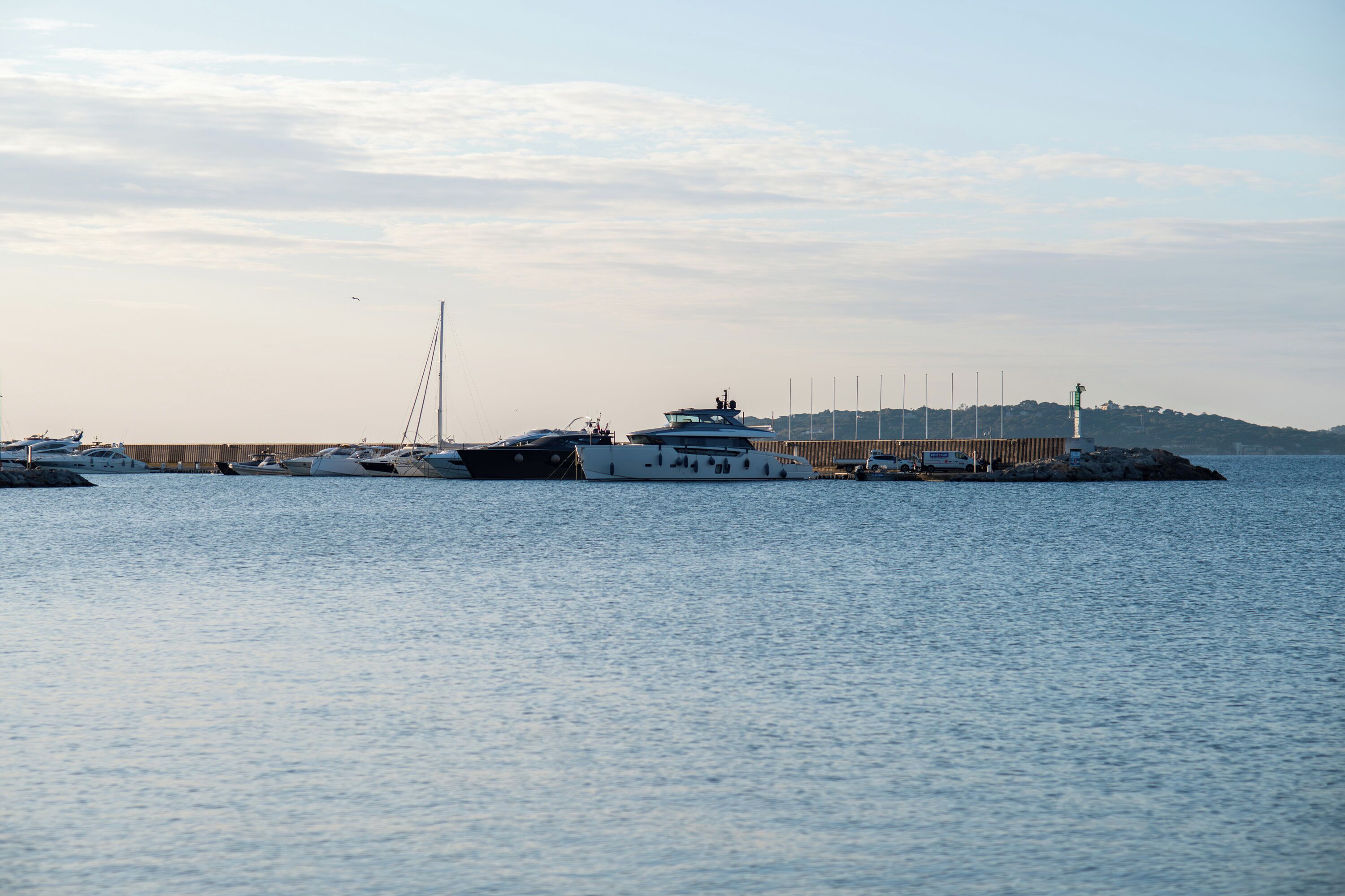 A marina with several yachts docked, a pier extending into the water, and a lighthouse in the background.