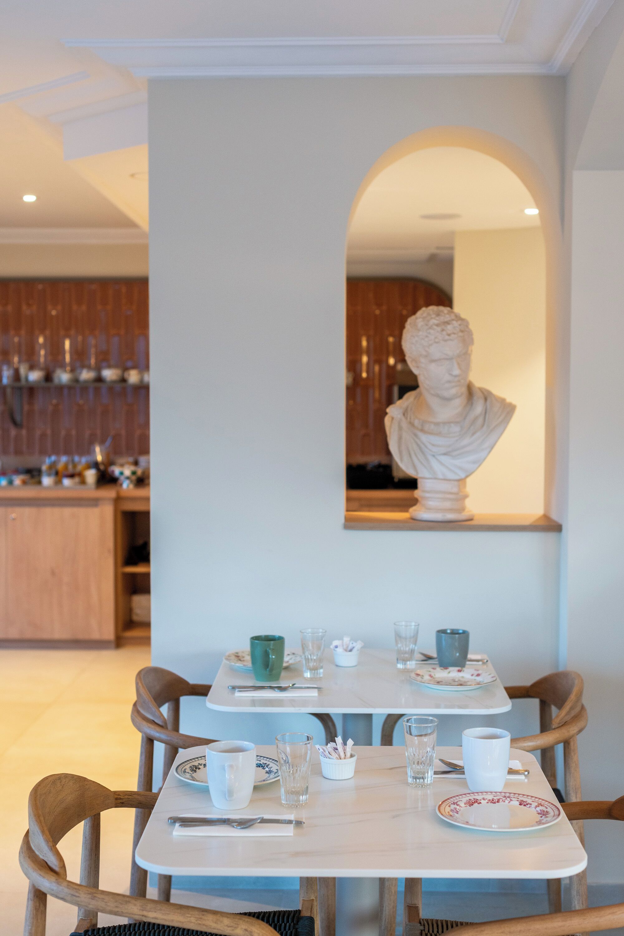 Un comedor con una mesa dispuesta para dos personas, un busto en la pared y una cocina al fondo.