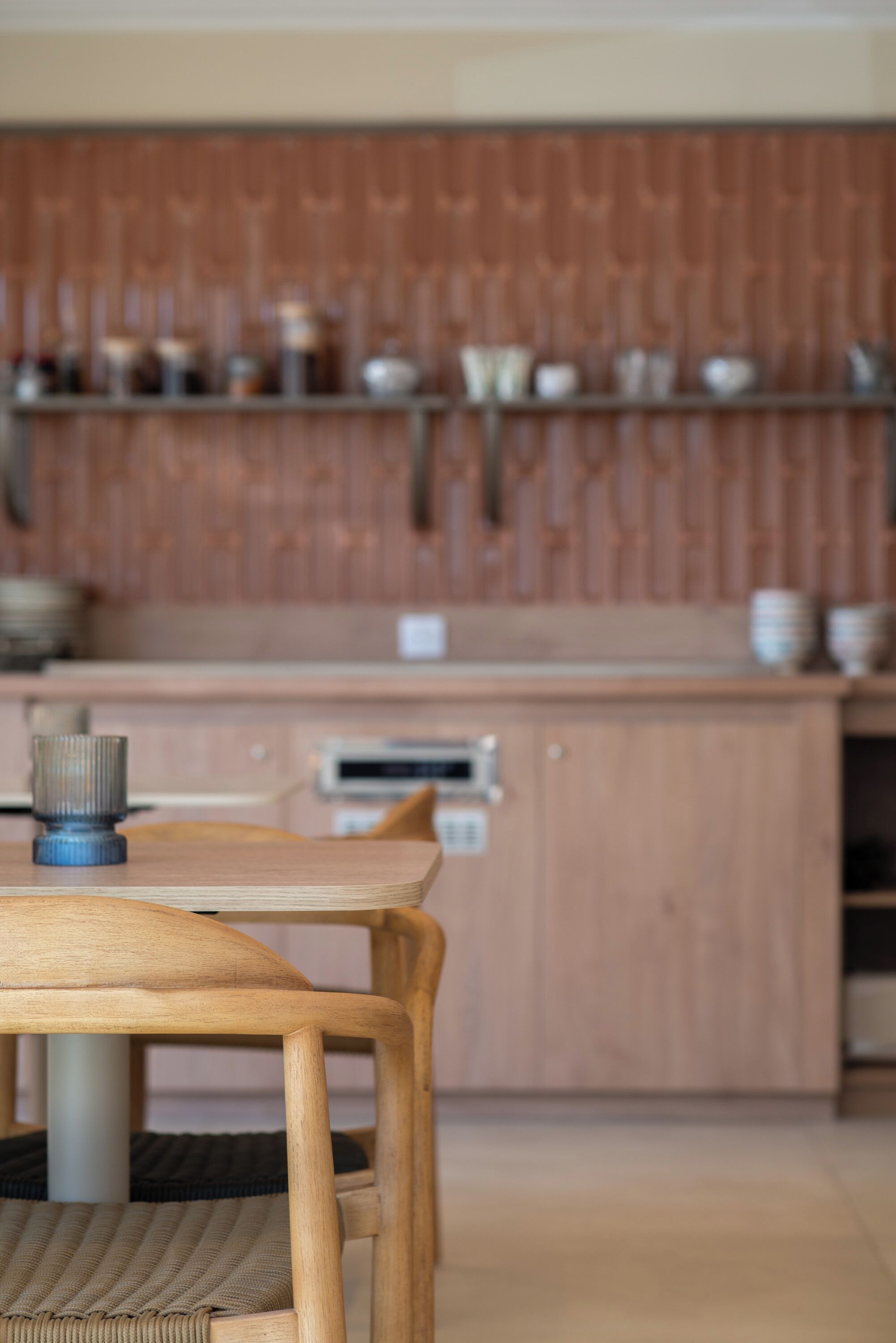 Una mesa de comedor de madera con sillas, una cocina con armarios y electrodomésticos, y una pared de madera con estantes que contienen vajilla.