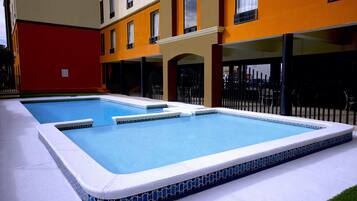 Una piscina rectangular con borde de azulejos, rodeada por un edificio de fachada roja y amarilla.
