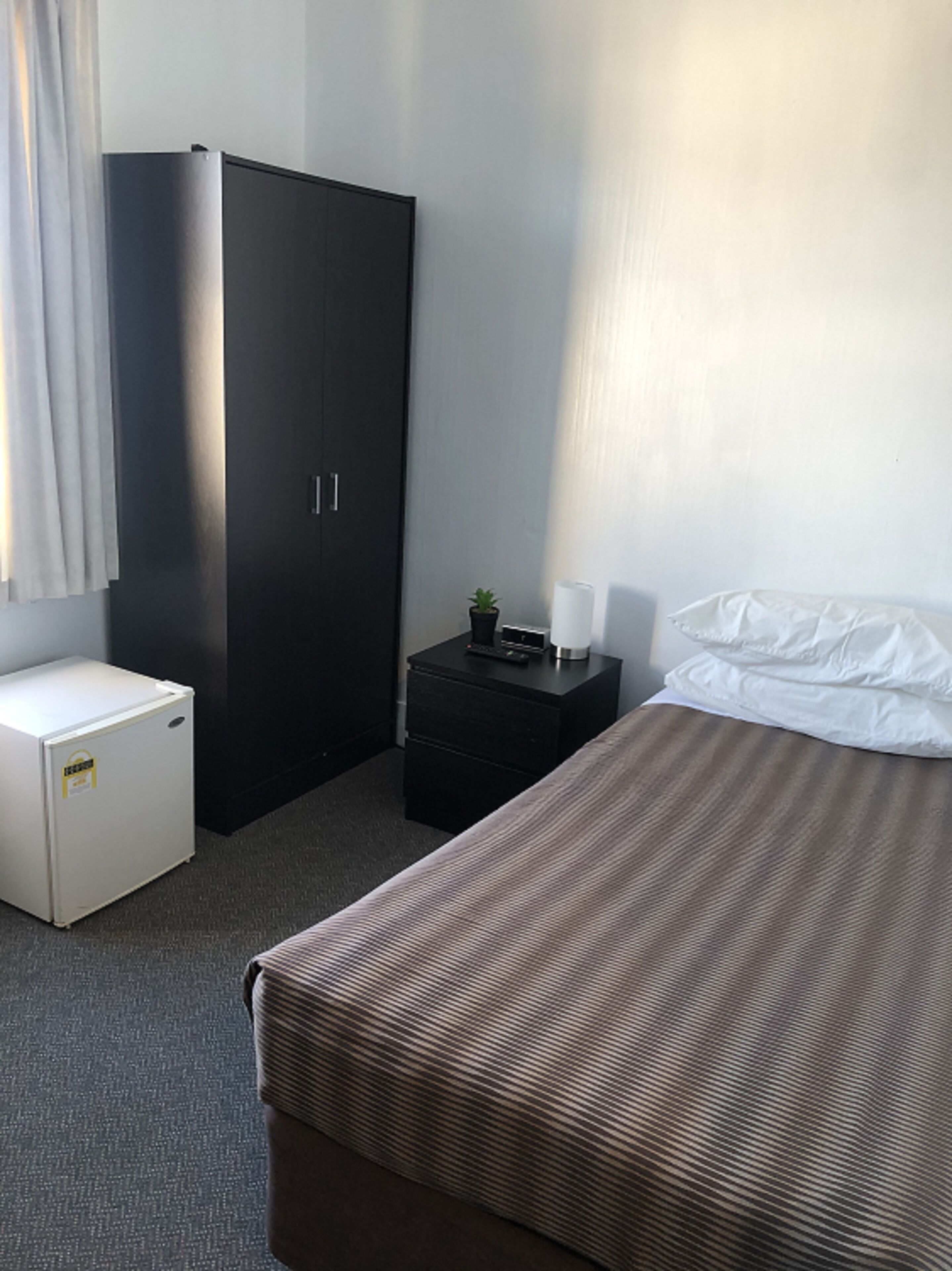 A hotel room with a bed, a wardrobe, a small desk with a lamp, and a refrigerator.