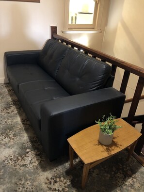 Un sofá de cuero negro con patas de madera, una mesita de madera con una planta en maceta y una ventana con cortinas.