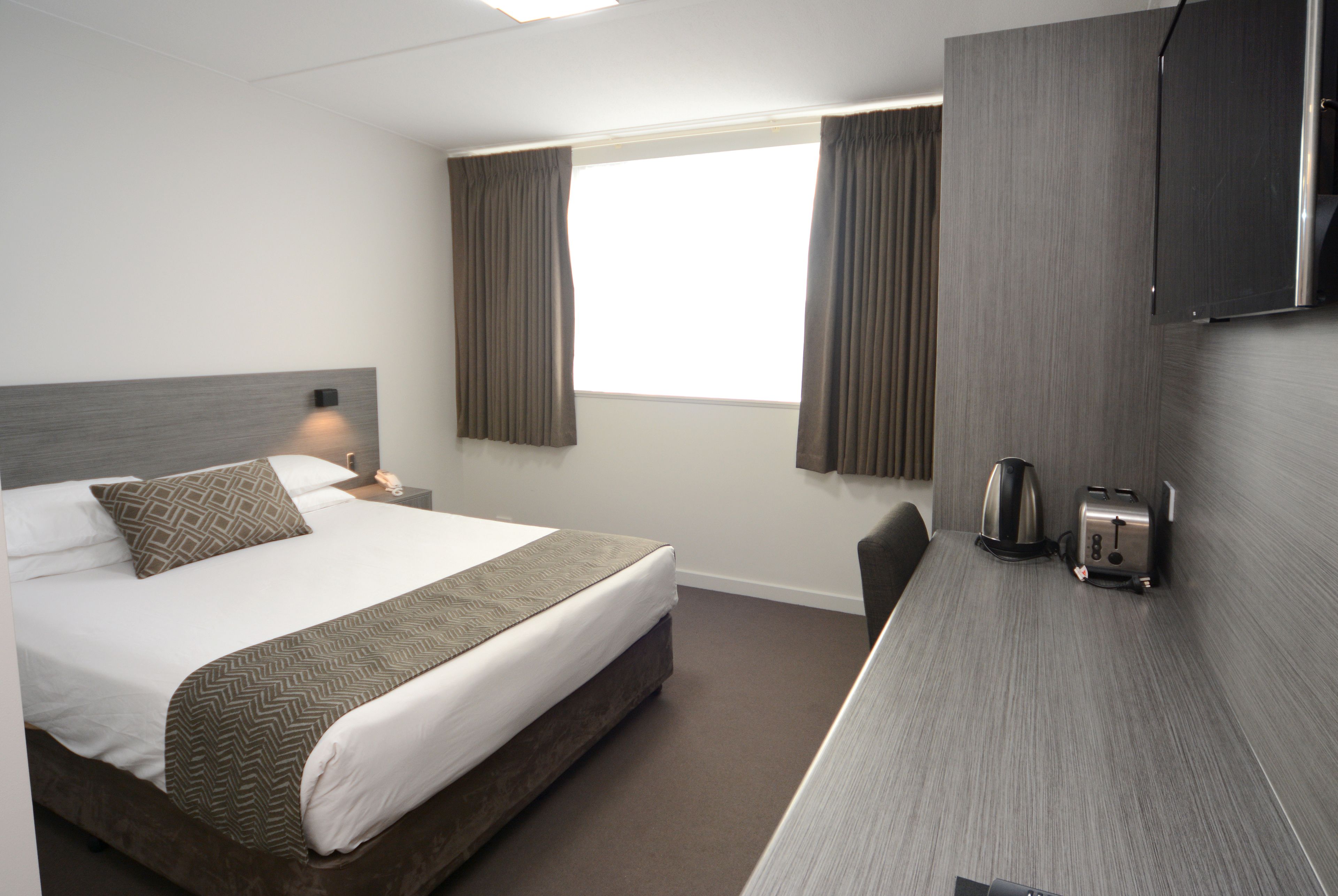 A hotel room with a bed, a desk with a kettle and toaster, and curtains on the window.