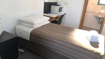 A single bed with a brown striped bedspread and white pillows, a small bedside table with a lamp, a microwave on a countertop, and a sink in the bathroom.