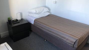 A small, single-bed room with a dark brown bedspread, a white pillow, a black nightstand with a small plant and a digital clock, and a grey carpeted floor.
