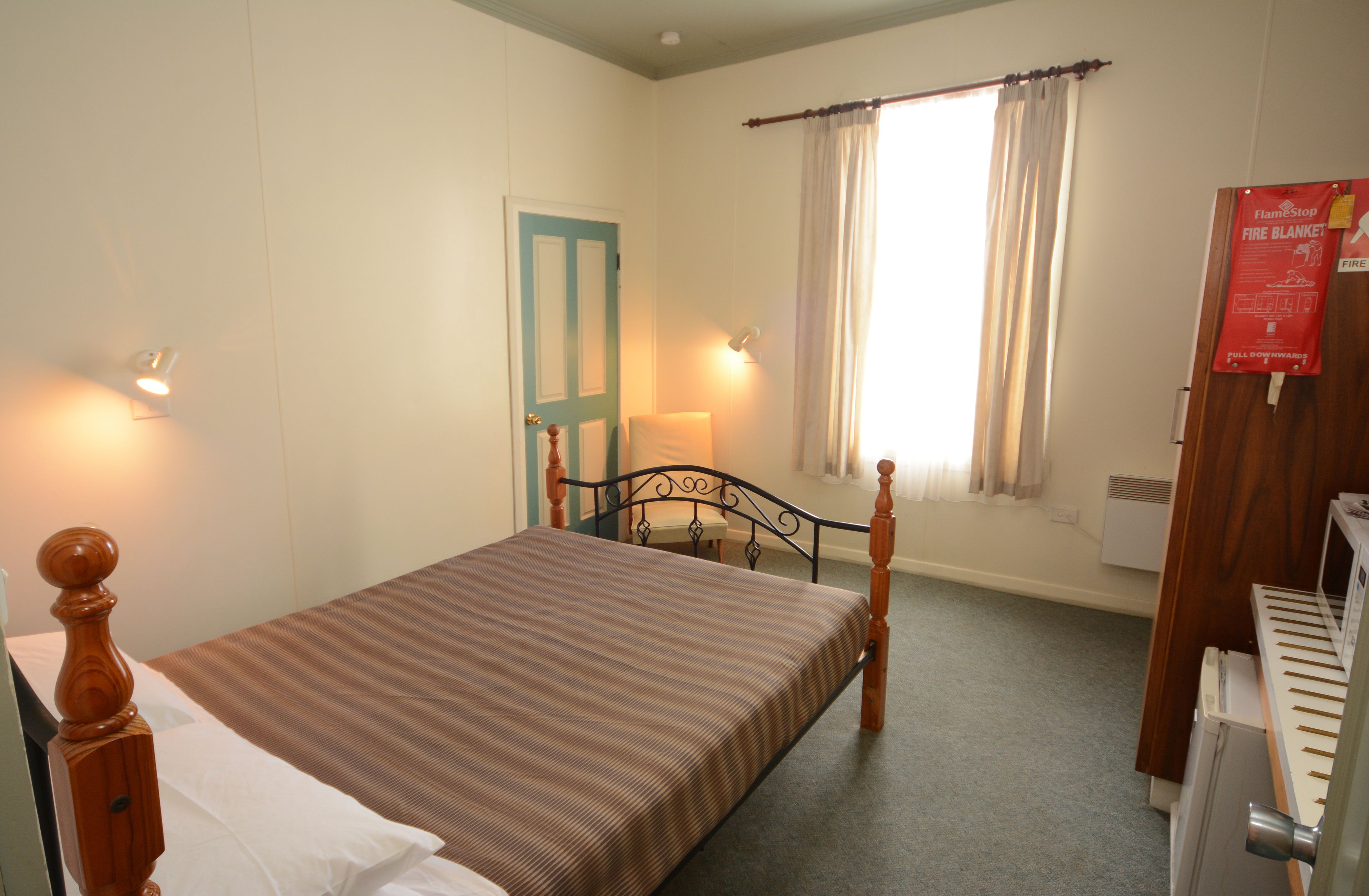 A bedroom with a bed, a window with curtains, a door, a wall-mounted lamp, and a radiator.