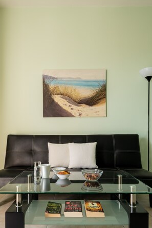 A living room with a glass coffee table, a black leather sofa, a painting of a beach, and a floor lamp. - Moonlight Apartments & Suites (Santorini)