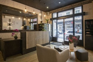 A hotel reception area with a wooden reception desk, seating, and a view of the street through large windows. - Hotel Ostende (Nice)