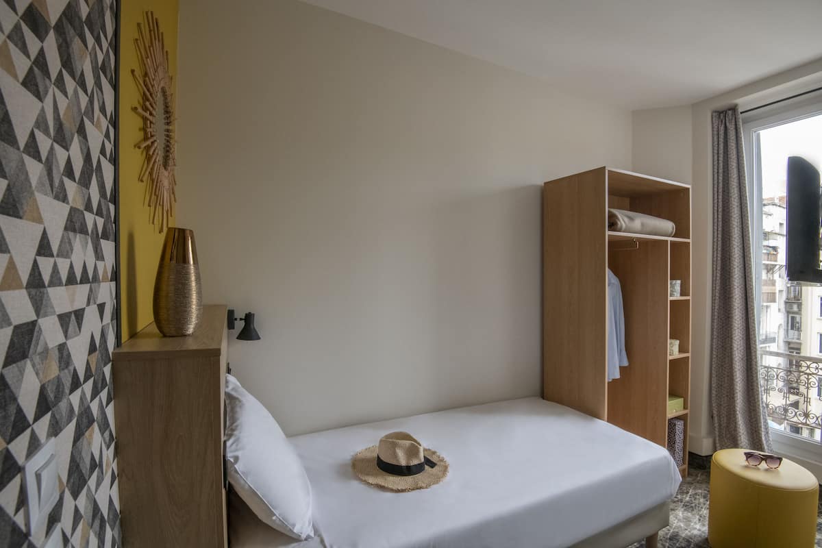 A hotel room with a bed, a wardrobe, a bedside table with a hat, and a window with a view of buildings.