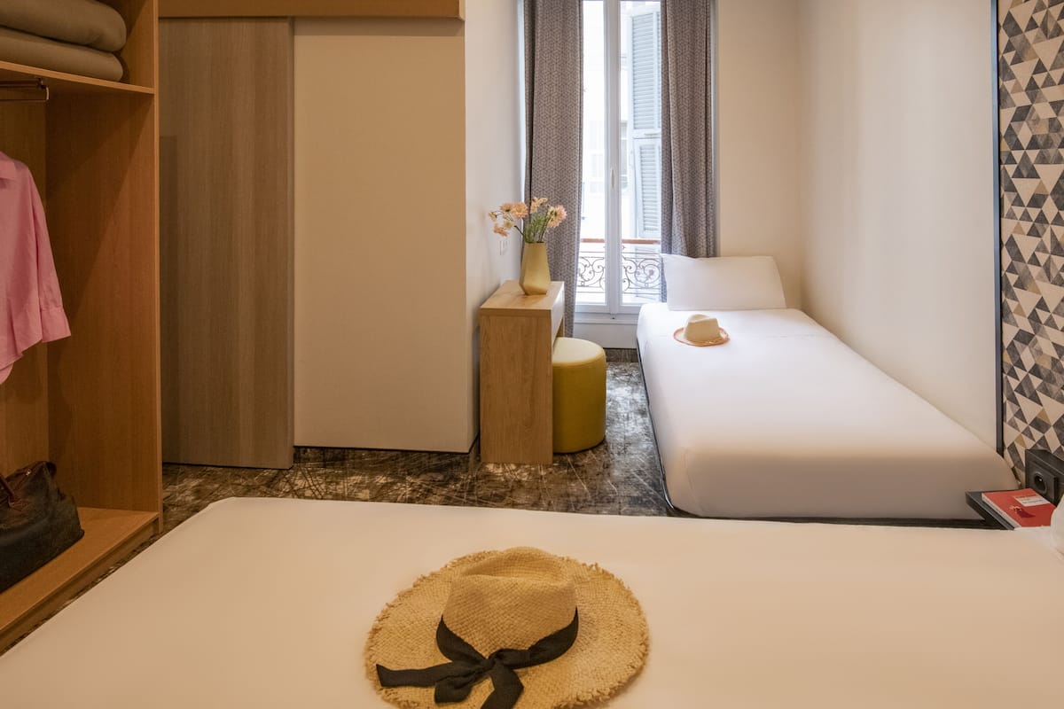 A hotel room with a bed, a small sofa, a wardrobe, a vanity table with a vase of flowers, and a window with curtains.