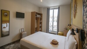 Habitación de hotel con cama, maleta, armario, televisor, espejo y ventana con vista a edificios.