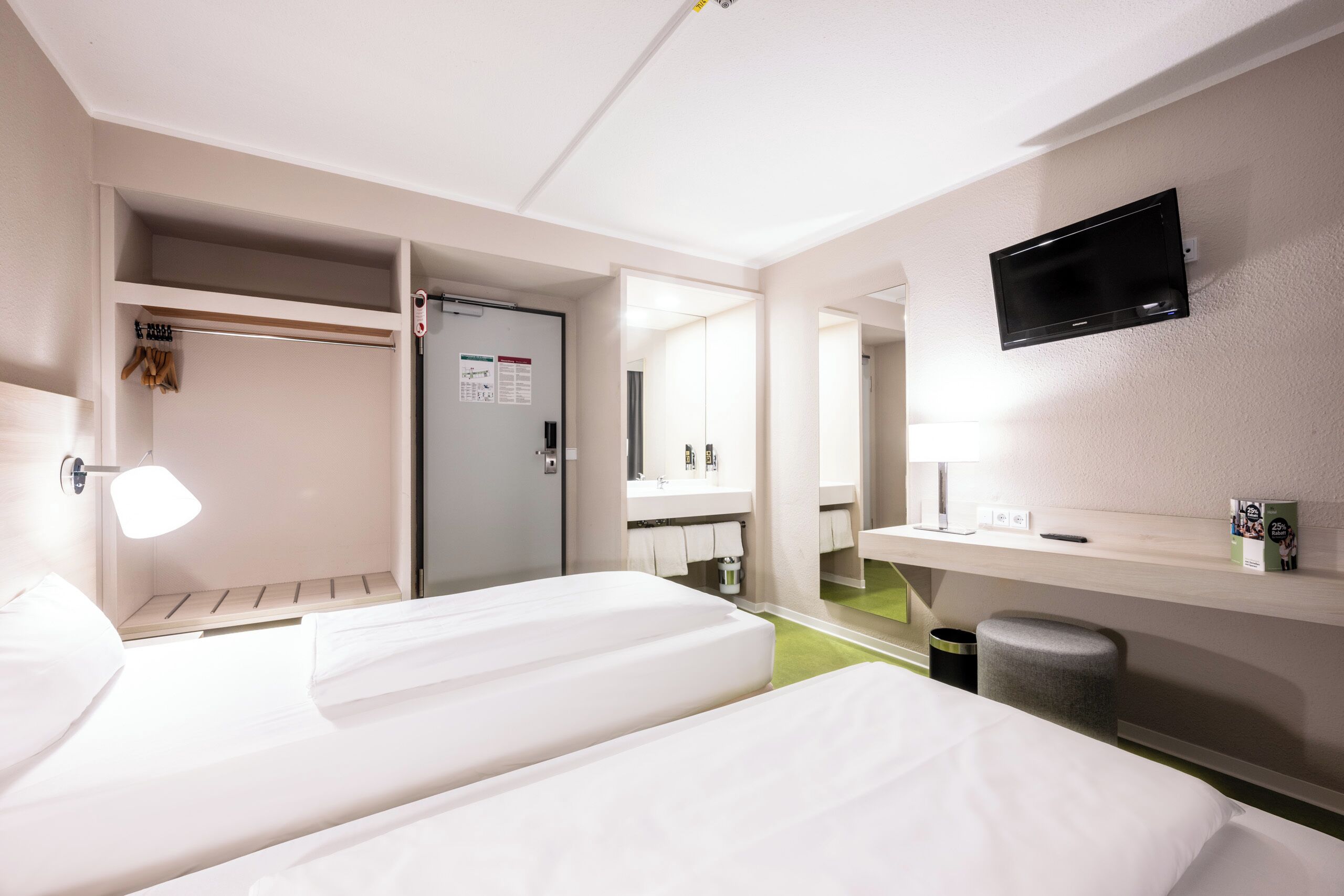 Ein Hotelzimmer mit zwei Betten, einem an der Wand montierten Fernseher, einem Spiegel, einem kleinen Schreibtisch und einem Papierkorb.