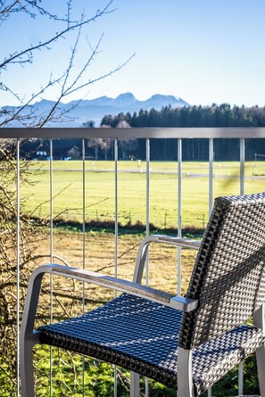 A wicker chair with a metal frame on a balcony overlooking a grassy field and mountains. - Coffee Fellows Hotel Irschenberg Süd (Irschenberg)