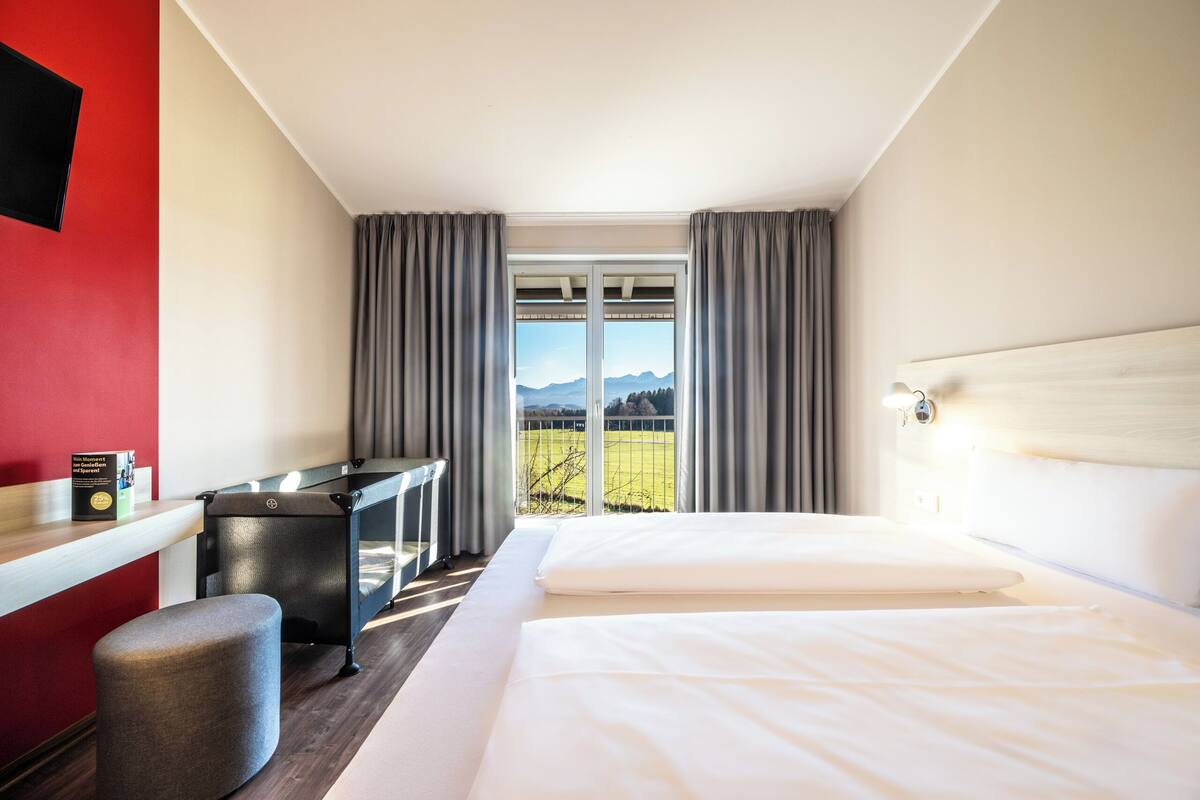 A hotel room with a bed, a desk, and a view of a green landscape through a large window.